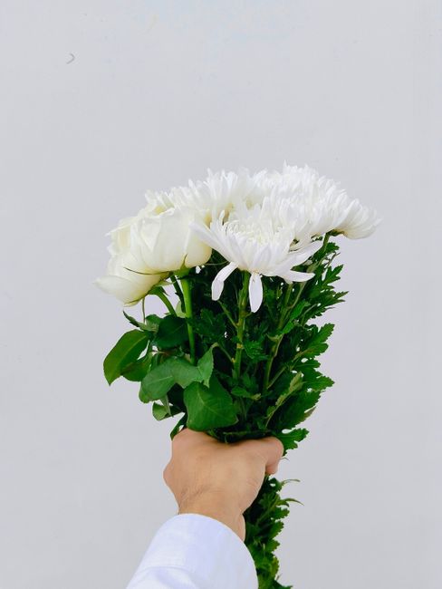 A hand holding a fresh floral bouquet featuring white chrysanthemums and roses, with lush green foliage and stems. The arrangement is wrapped in transparent cellophane, highlighting the vibrant white
