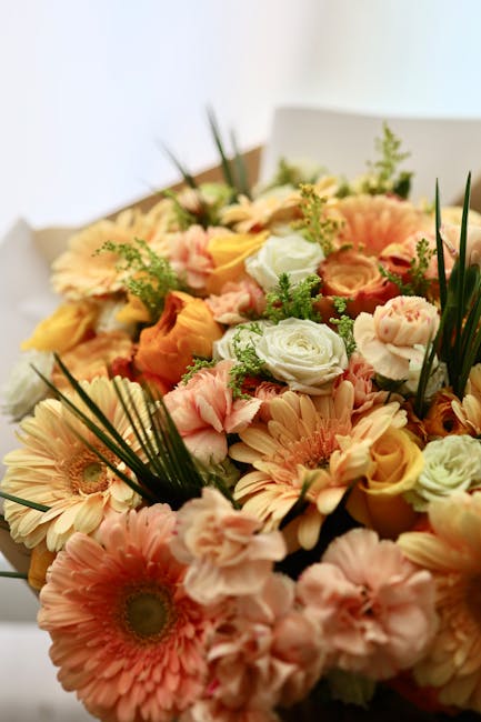 A close-up of a vibrant floral bouquet arranged with a variety of fresh flowers, including soft pink gerbera daisies, pale yellow roses, and white roses, complemented by sprigs of greenery and long, s