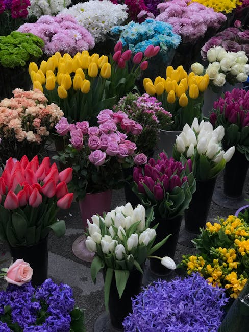 A vibrant display of various fresh flowers including yellow tulips with smooth petals and long green stems, light purple roses with tightly wound petals, pure white tulips with sleek, elongated blooms