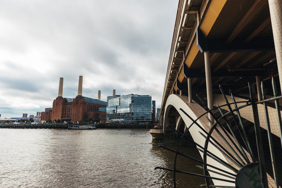 A view of the Thames River with modern buildings and industrial structures in the background, including the unmistakable red brick Battersea Power Station with four iconic chimneys. In the foreground,