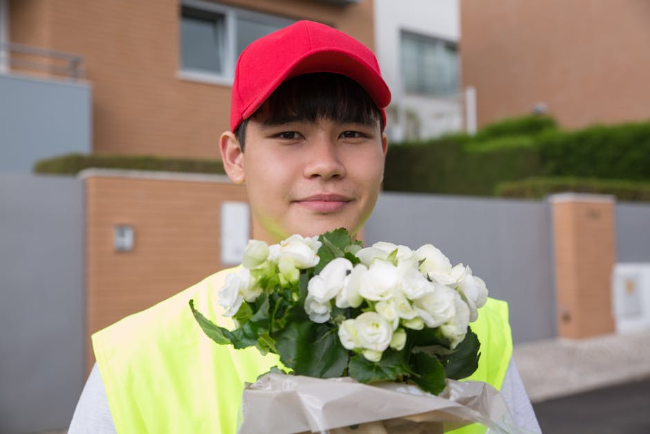 A young male delivery person wearing a bright yellow vest and red cap is standing outdoors in a residential area with modern brick and concrete houses, holding a bouquet of fresh white flowers wrapped