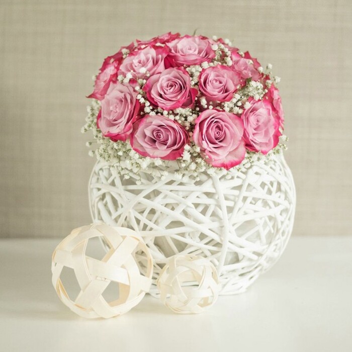 An elegant still-life arrangement presents a rounded bouquet of pink roses and white baby's breath standing proudly in a modern white woven vase, styled in a way that feels right at home in a chic Nine Elms riverside flat. The roses are the main attraction, gathered closely together so their heads form a soft, almost spherical dome. Each bloom shows a beautiful gradient of colour: the inner petals hold a shy, pale blush, while the outer edges deepen into a stronger, more saturated pink that borders on fuchsia, creating gentle contrast and depth. Threaded throughout the roses are countless tiny blossoms of white gypsophila, their fine stems disappearing between the petals so that only flecks of white seem to float in the bouquet like small, airy stars. The flowers are arranged in a spherical vase crafted from criss-crossing white strands, giving the impression of wicker or rattan painted in a pure, clean white. Its open lattice allows light to slip through, casting small, subtle shadows on the smooth white surface beneath. To the left of the vase, two decorative spheres made from thin, pale wooden strips echo the woven aesthetic, their loose geometric shapes and different sizes adding rhythm to the composition. Everything rests on a bright, unblemished white tabletop, which catches and softly reflects the light, heightening the sense of cleanliness and calm. The background is a gently textured wall in warm beige or cream, neutral and understated, almost like the softly painted walls of a new-build apartment looking out over Battersea Power Station and the wider Nine Elms area. The overall effect is one of tranquility and refined romance - a centrepiece suited to intimate celebrations, thoughtful gifts, or simply bringing a touch of softness to a minimalist London home.