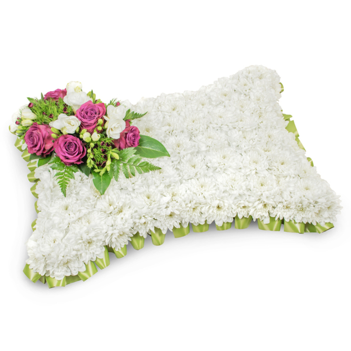 A tenderly made pillow-shaped sympathy arrangement, its entire rectangular form covered in a compact field of small, immaculate white chrysanthemums that create a smooth, plush surface reminiscent of a well-loved cushion. Each bloom presses close to the next, producing a uniform carpet of white that suggests peace, purity, and repose. Punctuating that serene expanse is a carefully placed accent on the upper-left corner: several deep fuchsia or magenta roses, their petals unfurling with quiet dignity, set among small white freesia-like blooms that add delicacy and a soft, floral lift. The accent rests on a halo of fresh greenery - slender fern fronds and assorted glossy leaves - which both supports the focal flowers and provides a verdant contrast to the white field. A light green satin ribbon, pleated or looped, traces the pillow's perimeter and subtly peeks out beneath the chrysanthemums, offering a refined border that echoes the natural foliage tones. Photographed against a crisp white backdrop with soft, natural shadowing, the arrangement's texture and three-dimensional form are clearly visible, carrying a mood of respectful remembrance. As a local florist familiar with Nine Elms, New Covent Garden Market and the riverside chapels near Chelsea Bridge, I often recommend this classic pillow for funeral services and graveside commemorations; its soft palette and understated elegance provide a gentle, comforting way to honour a life and offer solace to those who gather.