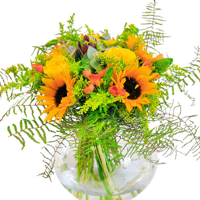 Radiating sunlit exuberance, this vibrant bouquet is thoughtfully arranged by a Nine Elms florist and offers a celebration of the season's brightest hues. Dominant sunflowers, their petals aglow in shades of deep gold and yellow, stand out like miniature suns, invoking joyful afternoons spent along the riverside walkways. At their side, buttery yellow roses unfurl in layers, each bloom meticulously curated to evoke classic garden charm. Crimson Hypericum berries nestle between the blossoms, their brilliant red orbs lending contrast and vibrancy, reminiscent of the lively produce at New Covent Garden Market. A lush gathering of greenery anchors the composition-the soft, feathery sweep of ferns, delicate sprigs of green filler speckled with dainty yellow blossoms, and slender flower spikes all blend harmoniously, creating movement and texture. Blue-green eucalyptus leaves peek out, adding a hint of aromatic freshness that recalls breezes flowing through local parks. The bouquet is set in a clear, round glass vase, its clarity emphasizing the structured tangle of fresh stems and water for a touch of urban elegance. As sunlight spills across the arrangement, it becomes a statement piece-perfectly suited for bringing warmth to a home, marking a milestone, or offering a heartfelt gift for friends and family in Nine Elms. Each element is selected with a florist's care, echoing the vibrancy and community spirit of the area.