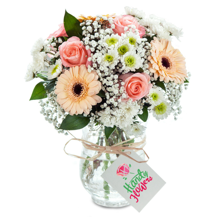 Set against a pure white background that mirrors the clean lines and modernity of Nine Elms, this carefully curated bouquet exudes both classic charm and contemporary flair. At the center, lush peach-pink roses spiral open in soft, inviting layers, their romance complemented by two vibrant gerbera daisies-each a stunning blend of peach and orange, with dark centers that command attention. White baby's breath fills the spaces, creating an airy, cloudlike texture, while clusters of crisp chrysanthemums, highlighted by lime-green centers, weave throughout, adding liveliness and visual contrast. Eucalyptus sprigs introduce a subtle woodland aroma and a graceful green accent, mirroring the park spaces and walkways along the Power Station area. Arranged in a clear, subtly ribbed glass vase through which healthy green stems and pure water shine, the bouquet is adorned at the neck with earthy twine and a bright white gift tag suitable for marking any occasion. The arrangement reflects the new energy of Nine Elms-versatile, cheerful, and thoughtfully crafted by local specialists-making it suitable for commemorating new beginnings, expressing gratitude, or simply celebrating a fresh season.