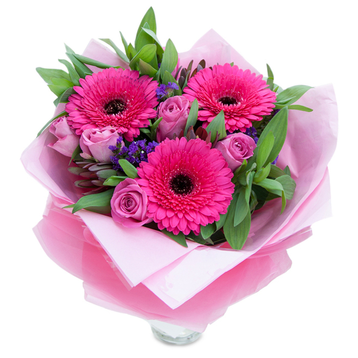 There is a playful elegance to this pink-dominant bouquet that feels both garden-fresh and carefully edited. Three fuchsia Gerbera daisies command attention with their expansive petals and dark centres, a vivid visual drumbeat around which softer elements gather. The roses present a quieter, dusty lavender-pink, their petals forming close, affectionate spirals that suggest tenderness and memory. Interspersed among them are little islands of purple statice, lending an airy, textural punctuation that makes the bouquet read as layered and alive. A generous spray of green foliage - broad salal or eucalyptus leaves with a faint, clean scent - gives the composition volume and a cool, verdant counterpoint to the warm pinks.
Wrapped in an inner cushion of matte pale pink tissue and an outer sheen of translucent fuchsia-pink cellophane, the bouquet is finished with folds that both protect and flatter the blooms; the wrap's shimmer subtly amplifies the pink tones. The clear glass vase rim peeking beneath the wrap suggests immediacy - this is a bouquet ready to sit on a kitchen table, a bedside chest, or a small dining table beneath the soft light that filters across Nine Elms Park. The scent is gentle: roses offering a soft, familiar perfume while gerberas carry a light, green freshness.
This piece is suited to intimate celebrations - birthdays, anniversaries, or a thoughtful congratulatory gift - and carries the quiet assurance of quality you'd expect from someone who shops at New Covent Garden Market and knows the neighbourhood's rhythms. It feels like an embrace in floral form, warm and reassuring.