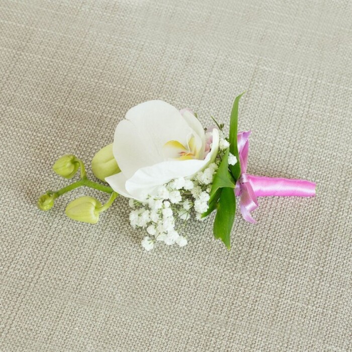 Displayed carefully on a gently textured, oatmeal-toned linen surface, this floral boutonniere feels like a small work of art crafted for a meaningful celebration in Nine Elms. The fabric backdrop, with its subtle weave and warm neutral shade, quietly frames the arrangement without distraction. At the centre lies a large, pristine white Phalaenopsis orchid, its broad petals unfolding in soft arcs. The petals appear almost luminous against the cloth, their velvety surface catching the light, while the orchid's throat reveals delicate details in soft yellow and faint blush pink, lending depth and a hint of warmth. Surrounding the base of the orchid, tiny tufts of white baby's breath form an airy cushion, each Gypsophila sprig composed of numerous miniature blooms that create a mist-like, romantic texture. A handful of slender, vibrant green leaves peek out from behind, providing crisp, fresh contrast that recalls the foliage seen in local gardens and walkways running through the heart of Nine Elms. Extending to one side, a short, branching stem displays two unopened orchid buds, their smooth, lime-green forms tightly closed, hinting at flowers that may open as the evening unfolds. The stems of the entire composition are meticulously bound in a bright fuchsia pink satin ribbon, the material glossy and smooth, wrapped snugly before being tied into a small, symmetrical bow whose tails lie neatly along the fabric. Gentle, even lighting bathes the scene, accentuating the refined textures of petals, baby's breath, and ribbon without casting hard shadows. The overall impression is one of classic elegance and thoughtful craftsmanship, a timeless boutonniere perfect for a groom, prom date, or special guest arriving at a ceremony near the Thames.