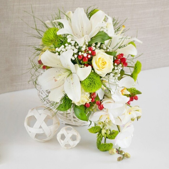 A meticulously styled white and green floral centerpiece rests on a smooth white surface, the kind you might find adorning a refined wedding table in a Nine Elms hotel overlooking the Thames. The flowers are cradled in a low, round basket woven from white wicker, its interlaced strands echoing natural craftsmanship and subtly reflecting the contemporary architecture of the area. Rising above the rim, several grand white Oriental lilies open wide, their trumpet-shaped flowers showcasing delicate dark freckles and golden anthers dusted with pollen, suggesting a rich, heady fragrance. At the centre, slightly elevated, a single creamy ivory rose blooms in perfect symmetry, its petals forming a soft spiral that draws the gaze and adds a hint of warmth to the cool white palette. Around these focal blooms, a generous selection of foliage fills the space: feathery green fronds, slender grasses, and glossy larger leaves providing a layered, almost garden-like structure. Among the greenery sit bright green, spherical dianthus or pom-pom chrysanthemums, their velvety, moss-like texture giving visual depth and interest. Fine clouds of white baby's breath thread delicately through the design, lightening the composition and adding a romantic haze. From the right side of the arrangement, a dramatic waterfall of white phalaenopsis orchids cascades downward, each flower with broad, glistening petals and pale yellow centres, creating a soft teardrop silhouette. Interspersed throughout the bouquet, clusters of rich red berries-likely hypericum-offer jewel-bright accents that instantly catch the eye and evoke the warmth of seasonal celebrations. To the left, two white decorative orbs, crafted from interwoven strands, rest on the tabletop, reinforcing the basket's texture and adding sculptural interest. A gently textured beige backdrop frames the entire scene, ensuring the floral artistry stands out, making this design ideal for wedding receptions, anniversaries, or elegant dinner parties across Nine Elms, Battersea, and the wider SW8 area.