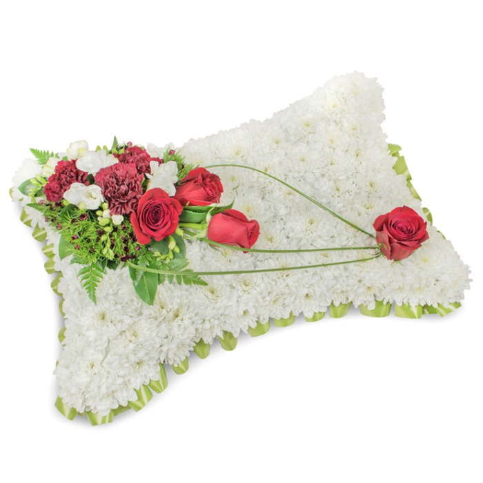 A lovingly composed rectangular memorial cushion made from a dense carpet of small white chrysanthemums, every bloom placed to create a plush, velvety surface that evokes purity and calm. Around the slightly raised perimeter a pale green satin ribbon peeks through, creating a subtle border that finishes the pillow with a touch of refinement. On the top left a clustered spray features deep, ruby-red roses alongside darker red carnations; their rich, layered petals are offset by small clusters of white freesias and fine, bright-green bear-grass, lending both fragrance and graceful line. Behind the flowers, soft fern fronds and glossy leaves provide a verdant backdrop that makes the reds sing against the white chrysanthemum base. From the main spray, three red roses trace a gentle diagonal toward the lower right, connected by thin, arching blades of grass that appear to hold a single perfect red rose at the pillow's end - a small, poignant focal point suggesting steadfast affection. The overall impression is one of serene dignity: the whites read as repose, the reds as remembrance, and the greens as a quiet echo of life. In soft daylight, the textures read clearly - the tiny chrysanthemum petals like fleece, the rose petals velvety to the eye, and the grass blades creating delicate arcs. Prepared with care for memorials and funeral services in the Nine Elms area - including delivery options across Nine Elms, Vauxhall and nearby Battersea Power Station - this arrangement is a classic expression of sympathy, offered by Florist Nine Elms with the discretion and craft expected for commemorations and church tributes.