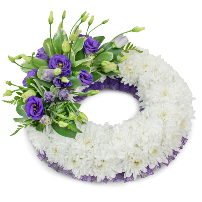Set against a pure white field, this circular memorial wreath reads as a composed testament to care and remembrance. The dominant surface is a compact ring of white cushion chrysanthemums - countless small, ruffled petals assembled to create a plush, uninterrupted circumference that feels tactilely soft and visually serene. Anchoring the design in the upper-left quarter is a lush accent of purple Eustoma (lisianthus), where rich violet flowers sit beside paler lavender varieties and slim buds, offering a pleasing gradient of purple hues and a supple, velvety texture. Greenery - a blend of broad, lightly variegated leaves and slender, fern-like fronds - surrounds and supports the lisianthus, providing contrast, botanical depth and a breath of living green. Discreetly visible beneath the chrysanthemums is a neat trim of dark purple mesh or lace, its crinkled weave tracing both the internal and external edges of the wreath and harmonising with the floral accent. The studio-style lighting produces a faint, diffused shadow that lifts the wreath from the white surface and reveals the arrangement's gentle volume. Picture this piece brought to a service near Vauxhall Bridge or laid beside graves in local cemeteries: it offers solemnity without excess, a clear language of sympathy where white signifies peace and purple gestures toward lasting remembrance and respect.