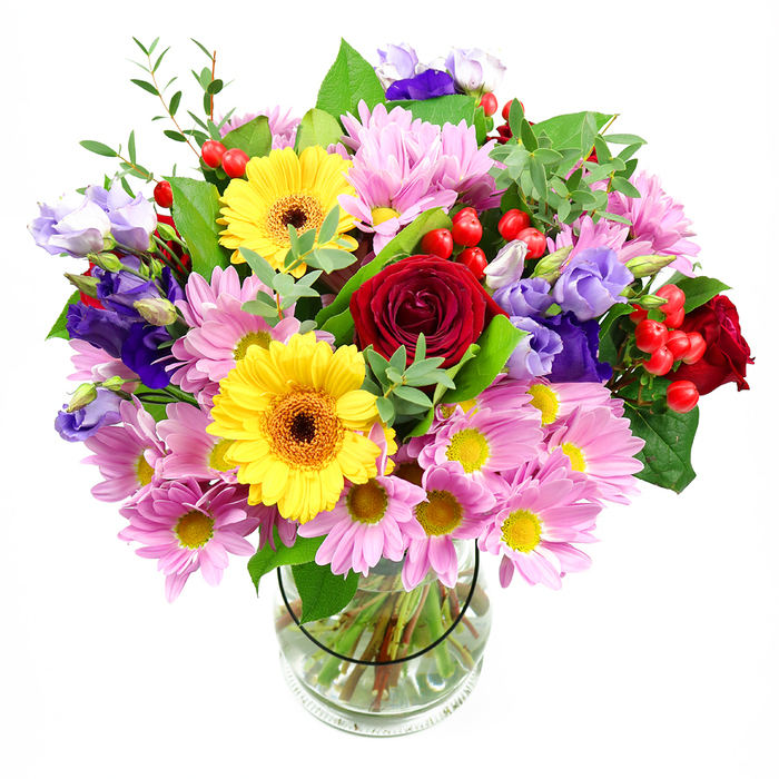 In this image, a lush, overflowing bouquet rises from a simple clear glass vase, the crisp white background allowing every shade of red, pink, yellow, and purple to glow. Deep crimson roses form the central cluster, their folds of velvety petals inviting the hand and eye to linger, giving the display a timeless, romantic presence. Nearby, two yellow gerbera daisies open wide like little suns, their golden petals radiating from warm brown-orange centres that add a confident, cheerful note. Surrounding these focal flowers is a generous ring of pink daisies and chrysanthemums, their many fine petals and petite yellow eyes creating a soft, cushion-like base that makes the arrangement feel both full and airy. Slender stems of purple and lavender lisianthus weave through this pastel cloud, their bell-shaped flowers and buds introducing cool, refined tones that bring balance and a slightly contemporary edge. Tucked between the blooms, rounded clusters of glossy red hypericum berries glint like tiny lanterns, offering visual rhythm and a pleasing, tactile contrast. The whole bouquet is laced with varied greenery-broad, dark leaves providing structure, while lighter green sprigs and shoots reach outward, giving the composition movement, as though it has a gentle breeze drifting through it. Through the transparent vase, the viewer can see an elegant network of stems immersed in clear water, testifying to careful conditioning and expert arranging. It feels like the kind of sophisticated yet welcoming bouquet a Nine Elms local might place by a balcony window overlooking the Thames Path, perfect for birthdays, housewarmings, or simply bringing a burst of natural colour into a modern London flat.