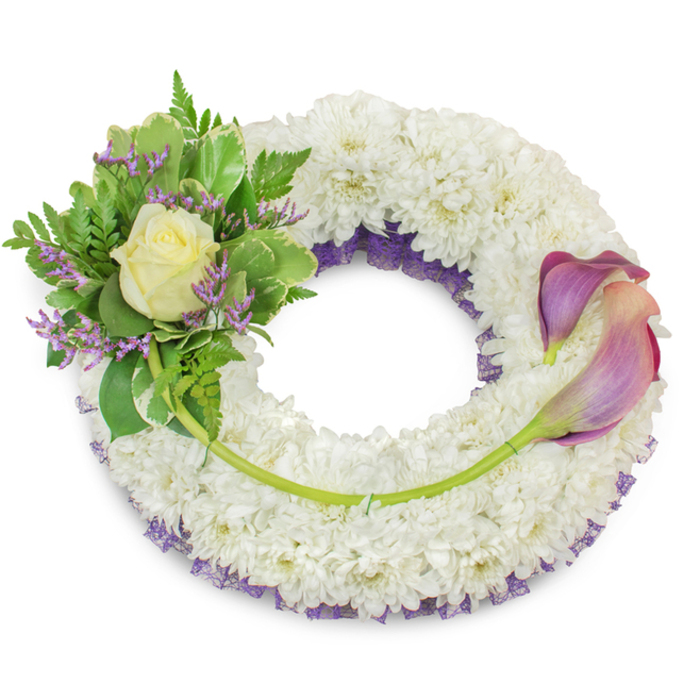 An elegant, full circular wreath photographed on a clean white field, where a thick ring of pristine white chrysanthemums forms a soft, cloud-like base that visually conveys peace and reverence. Beneath the white mass a deep purple ruffled netting peeks through around the inner edge, adding a refined layer of colour that anchors the design without detracting from the overall serenity. Arranged on the upper-left quadrant, a composed cluster centers on a single cream-tinged rose, its petals unfolding with quiet grace; surrounding it, broad variegated leaves with paler edges and fine fern fronds create a tapestry of greens, while tiny lavender-hued statice are scattered through the foliage to echo the hidden purple beneath. On the wreath's right side two graceful calla lilies extend from about two to five o'clock, their trumpet forms presenting a soft gradient from deeper mauve or purple at the tips to a gentle peach-pink nearer the base, their long green stems curving along the lower-right arc and visually connecting to the left cluster. A solitary longer stem travels from the left grouping across the lower wreath, anchored near the callas to provide a discreet line that guides the eye. The composition is calm and measured, the textures of chrysanthemum, rose, variegated foliage and delicate statice harmonising to offer a dignified sympathy piece. Imagined for memorials or graveside services in Nine Elms, Vauxhall and surrounding streets like Wandsworth Road and Nine Elms Lane, this wreath conveys respectful remembrance and enduring comfort.