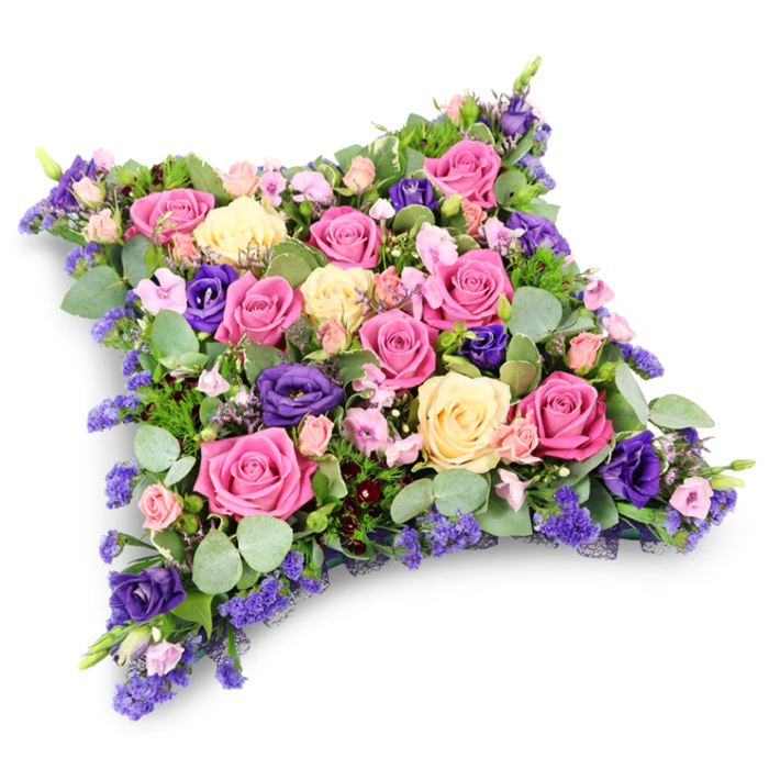 A detailed floral cushion tribute is shown against a bright, uncluttered white backdrop, its rectangular shape softened into a plush pillow of blossoms that seems to invite a moment of reflective stillness. The design is dominated by full-headed roses in complementary shades: vivid fuchsia and cerise at the heart of the composition, surrounded by softer pastel pinks and creamy ivory-yellow blooms. Each rose is carefully angled so that its layered petals open outward, forming a tapestry of ruffles and spirals that draw the gaze slowly across the arrangement. Interspersed with these classic roses are lush violet and royal purple lisianthus, their gently ruffled petals adding a velvety texture and a slightly cooler depth of colour, like pockets of evening shade among sunlit petals. Scattered throughout are sprays of tiny lavender, lilac, and blue-toned filler flowers, likely statice or limonium, which lend a delicate, almost airy finish to the dense composition. Around the edges and peeking between blossoms, rounded eucalyptus leaves in dusty sage green blend with slim, feathered fern-like foliage, creating a soft green frame that makes the pinks, creams, and purples appear even richer. The overall effect is of a lovingly constructed funeral pillow, full and generous yet serene, suitable for a farewell service in one of the local churches or crematoria serving the Nine Elms community. The tribute feels both modern and timeless, echoing the changing skyline along the Thames while preserving the gentle traditions of condolence flowers. It is the kind of carefully balanced design that a skilled Nine Elms florist would prepare for families who want their funeral flowers to convey respect, comfort, and lasting remembrance.