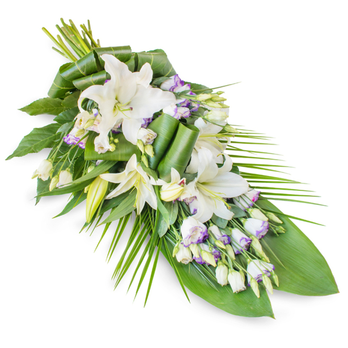 A classic teardrop funeral spray of white lilies and lisianthus is shown on a stark white background, its lush upper portion tapering smoothly into a slender, pointed tip, as though prepared to rest on a casket or memorial table in the Nine Elms area. The focal point is a group of white oriental lilies, some fully open and spreading their broad petals in serene, sculptural arcs, others standing tall as creamy buds that promise to open in the days following the service. Their throats glow with a soft green hue, while fine golden stamens add a gentle warmth amidst the cool whites. Interwoven among these stately blooms are clusters of lisianthus, their smaller, bell-like flowers offering a cloud of white petals delicately edged with hints of lavender and light purple, creating a soft, watercolour-like transition of shade. Tiny, unopened lisianthus buds and emerging blooms dot the arrangement, adding detail and the suggestion of continuity and ongoing love. A rich foundation of foliage supports and frames the florals: broad, dark green leaves create a deep, velvety background, while several leaves are folded, curled, and rolled into elegant, architectural ribbons that lend a contemporary edge. From the lower side, slender palm-style fronds sweep outward and down, echoing the gentle curve of the Thames paths near Embassy Gardens and guiding the eye along the length of the spray. The entire design exudes quiet formality and heartfelt respect, suitable for funeral services, crematorium ceremonies, and celebration-of-life gatherings across Nine Elms, expressing condolence, remembrance, and a wish for peaceful rest without a single word spoken aloud.