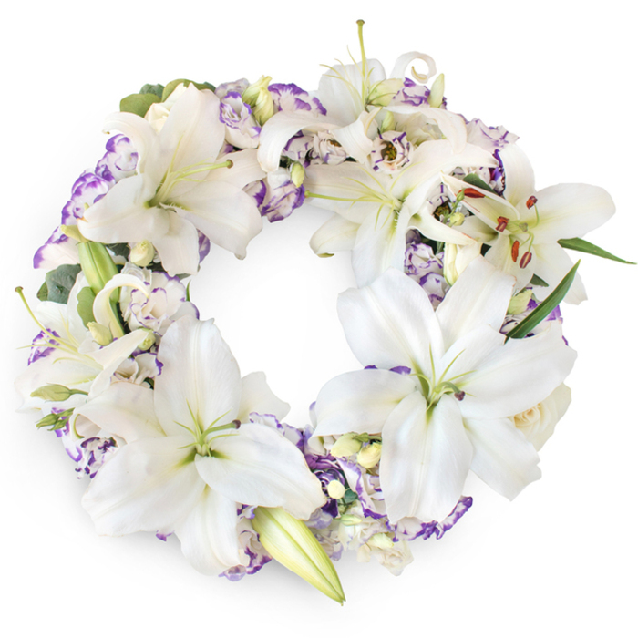 A full, circular funeral wreath of white lilies and purple-tinted blooms is shown against a crisp white background, every detail standing out with dignified simplicity. The design is abundant yet controlled, dominated by large, snow-white Oriental lilies that open outwards to form a halo of soft, star-shaped flowers. Each lily displays a pale green throat and delicate veins running through the petals, while central stamens carry touches of golden-brown pollen that add a quiet, natural warmth. Around and between these focal flowers, smaller lisianthus blooms unfurl in ruffled layers, their white petals outlined in shades of lavender and violet, bringing a gentle hint of colour rather than a bold contrast. Some flowers are fully open, others in tight buds, giving the wreath a natural rhythm, reminiscent of watching the seasons change along the paths of nearby Battersea Park or the evolving skyline of Nine Elms. The florist has tucked in sprigs of fresh greenery - perhaps slender leaves and eucalyptus-like stems - which emerge subtly from beneath the blossoms to provide structure and a cool green undertone. The circle is completely filled, with no gaps, evoking wholeness, eternity, and the unbroken connection between the departed and those who remain. The overall impression is calm, reverent, and tender, ideal for a memorial service, church funeral, or crematorium ceremony in and around Nine Elms. It feels like a carefully crafted tribute created by a local floral designer who understands how to express sympathy, love, and respect through pure white petals and soft lavender accents.