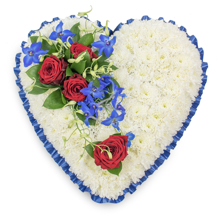 This image shows a carefully crafted heart-shaped funeral tribute designed with the calm, ordered beauty that many families in Nine Elms choose for a final farewell. The entire surface of the heart is densely packed with white chrysanthemum blooms, each flower head touching the next to create a seamless, plush layer of petals like fresh snow. Around the edge, a royal blue satin ribbon is folded into even pleats, hugging every curve of the heart and giving a crisp outline that stands out clearly against the background. The ribbon's sheen catches the light in soft highlights, lending the piece a feeling of quiet formality suitable for a church, crematorium, or graveside service. On the upper left, a focal spray of flowers rests diagonally across the chrysanthemums: three deep red roses, fully open, reveal their layered, velvety centres and bring a strong note of love and devotion. Dark green leaves cradle the roses, while bright electric blue dendrobium orchids extend downward from the cluster, their delicate, star-shaped blooms adding fluid lines and a modern colour accent. Light green, curling tendrils thread through the arrangement, softening edges and evoking natural growth. On the lower right curve of the heart, a single red rose is placed with intention, echoing the main group and gently drawing the eye across the design. The tribute has a serene, respectful presence, the white, red, and blue palette suggesting purity, affection, and peace-an arrangement a local florist near the Thames in Nine Elms might prepare to accompany condolences at a family gathering or chapel of rest.