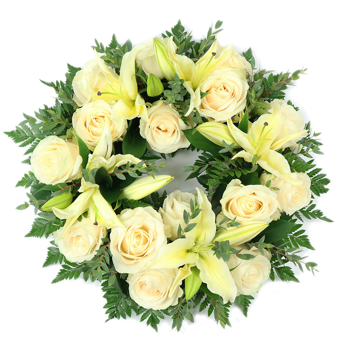 Photographed flat from above on a simple white surface, this circular funeral wreath presents a composed, harmonious blend of soft creams, pale yellow and verdant greens. Approximately fifteen to twenty fully opened creamy white roses form the principal visual weight, their layered, tightly spiralled petals catching a soft, even light that lends them a porcelain-like clarity. Interspersed deliberately among these roses are delicate lemon-hued lilies - some unfurled to reveal elegant trumpet shapes and subtle pale stamens, others held as poised buds offering textural variety and a sense of unfolding life. The blooms are set against a dense, deep-green base of foliage: fine, feathery fern fronds provide an airy, delicate perimeter while broader, rounded sprigs of eucalyptus or similar leaf add depth, cool tones and a gentle fragrance. The arrangement's dense packing and symmetrical balance give the wreath a full, luxurious presence without appearing heavy; instead it reads as refined and consoling. The circular form, precise and even, underscores themes of eternity and remembrance, making the piece appropriate for memorial services, graveside tributes or a quiet display in a funeral chapel. The combination of velvety rose surfaces, the lilies' smooth, curving petals and the textured greens creates a sensory whole that is simultaneously tactile and serene. Positioned for clarity against the plain background, the wreath communicates a respectful simplicity, a composed expression of sympathy suited to families in Nine Elms, Vauxhall and the neighbouring riverside districts.