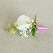 A single, carefully crafted boutonniere is laid horizontally on a softly textured, oatmeal-coloured linen cloth, the neutral fabric allowing every floral detail to stand out with quiet elegance. At the heart of the piece rests a pristine white Phalaenopsis orchid, its broad, velvety petals opening in a gentle curve. The orchid's centre glows with soft yellow and the faintest blush of pale pink, like a subtle sunrise caught within the bloom. Around the base of the orchid, tiny white clouds of baby's breath cluster together, each delicate Gypsophila floret adding an airy, romantic haze that softens the design. Slender, vibrant green leaves peek out behind the orchid, their smooth surfaces offering a fresh contrast to the snowy petals and giving the boutonniere a sense of natural movement, as if just gathered from a riverside walk near the Thames in Nine Elms. To one side, a short stem carries a pair of tightly closed, lime-green orchid buds, suggesting the quiet promise of flowers yet to open later in the day. All the stems are neatly bound with a bright fuchsia pink satin ribbon, the glossy bow tied with a florist's precise hands, its tails lying flat against the linen. The lighting is even and gentle, with no harsh shadows, highlighting the fine textures of each petal, the tiny spheres of baby's breath, and the subtle weave of the fabric. The overall impression is of refined simplicity and celebratory grace, perfect for a groom or prom guest stepping out from a Nine Elms townhouse for a special occasion.