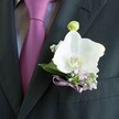 A close-up photograph shows a sharply tailored charcoal suit jacket worn over a pale grey shirt with a fine woven texture, the fabric catching soft indoor light. At the heart of the image is a richly coloured, neatly knotted tie in deep magenta, leaning towards plum, its silky sheen echoing the romantic mood of a wedding or black-tie event in Nine Elms. Pinned precisely to the left lapel, just above the breast pocket, is a white Phalaenopsis orchid boutonniere, crafted with the care of an experienced florist. The orchid's broad, velvety petals open like sculpted wings, flawless and luminous against the dark cloth, with a vivid yellow and soft orange centre adding warmth and depth. Surrounding the orchid, airy sprigs of baby's breath and petite lilac-toned blossoms create a cloud of gentle colour that softens the strong lines of the suit. A fresh green orchid bud rises just above the main bloom, its pale lime tone suggesting new beginnings and the quiet anticipation of the ceremony. Deep green foliage frames the flowers, its slightly glossy surface offering a natural counterpoint to the matte lapel. At the base of the arrangement, a slim lavender satin ribbon is tied into a precise bow, its sheen mirroring the tie and hinting at a carefully chosen colour palette that might match bridesmaid dresses or other wedding flowers. The overall impression is of understated elegance and personal detail, the kind of lapel flower a groom might wear as he walks through the riverside streets of Nine Elms towards his vows, carrying with him a subtle, fresh scent and the calm confidence of a thoughtfully planned day.