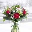 A luxurious red and white bouquet stands proudly in a clear cylindrical glass vase, its stems freshly cut and visible through the clean water, like a little piece of Nine Elms sunshine brought indoors. Deep red roses form the heart of the arrangement, their velvety petals unfolding in measured spirals that feel both confident and romantic, while a large ruffled crimson carnation at the front adds bold texture and drama. Around them, soft white alstroemeria and other creamy blossoms open gently, their fine speckles and curved petals creating a feeling of purity and calm that balances the passionate reds. Tightly closed white lily buds rise above the cluster, promising future blooms and lending a quiet sense of anticipation, as if the bouquet will continue to change and delight over several days on a table overlooking the Thames or a bright kitchen near Battersea Power Station. Sprigs of delicate white gypsophila, or baby's breath, float between the larger flowers like tiny stars, giving an airy, cloudlike fullness. The blooms are cradled by glossy, dark green tropical leaves that are carefully curled and wrapped around the base, forming a sculpted collar that hints at professional care and attention. Additional layers of eucalyptus-like sprigs and pointed green foliage introduce cooler tones of green, adding depth, movement, and a faint woodland freshness to the composition. Resting on a spotless white surface against a softly blurred, bright background, the bouquet is bathed in even natural light, making each colour appear crisp and uplifting. The overall impression is of thoughtful celebration and warm affection, ideal for an anniversary dinner in a Nine Elms riverside apartment, a heartfelt birthday surprise, or a refined welcome arrangement in a local office reception.