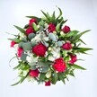 Viewed directly from above against a clean white background, this abundant hand-tied bouquet forms a near-perfect circle of colour and texture, as if it has just been set down on a florist's bench in Nine Elms. At its heart, velvety red roses in varying stages of bloom draw the eye, some still in tight buds, others gently unfurling to reveal deep crimson petals that suggest romance and timeless affection. Woven between them are ruffled carnations in vivid fuchsia and ruby red, their layered petals creating a soft, feathered look that feels joyful and celebratory. Clusters of dainty white spray chrysanthemums, each flower like a tiny daisy, bring light into the arrangement, adding a fresh, cheerful brightness that lifts the richer tones. Threaded delicately throughout is a cloud of fine white gypsophila, or baby's breath, its tiny blossoms forming a misty veil that softens every edge. Around the blooms, lush greenery builds a generous frame. Dark, pointed leaves, likely from lily or alstroemeria stems, create structure and a sense of movement, while rounded eucalyptus leaves in soft silvery green add a contemporary twist, suggesting a gentle, refreshing fragrance. The lighting is even and natural, highlighting every curve of petal and vein of leaf, making the bouquet feel as if it has just been prepared for same-day delivery to a home overlooking the Thames or a new apartment near Battersea Power Station. This richly coloured arrangement radiates warmth and celebration, perfect for anniversaries, Valentine's surprises, or simply sending heartfelt love and encouragement across Nine Elms and the surrounding streets.