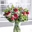 A lush, vibrant bouquet of deep crimson red roses and matching ruffled carnations stands proudly in a clear glass vase on a bright white tabletop, bathed in soft natural light from a large window typical of modern Nine Elms apartments. The roses range from tight, promising buds to fully open blooms with velvety petals curling outward, while the carnations add plush volume and a classic, almost nostalgic texture. Nestled between the bold reds are clusters of pristine white alstroemeria, some still in elegant bud form and others partially open, their trumpet-shaped petals edged with the palest green. Delicate sprays of tiny white gypsophila, or baby's breath, float throughout the design like a fine, cloudlike mist, softening the contrasts and giving the whole bouquet a romantic, airy feel. Surrounding the flowers, broad, glossy green leaves form a strong collar at the base, while slimmer, lighter green foliage threads upward, creating gentle movement and depth, as if the bouquet had just been gathered from a riverside garden along the Thames near Nine Elms. The stems are neatly cut and clearly visible through the water in the vase, reinforcing a sense of crisp freshness and professional care. Behind, the view through the window is softly blurred and almost monochrome, so the rich reds, cool whites, and layered greens appear even more vivid, commanding attention without feeling loud. The arrangement feels like a refined yet heartfelt gesture-perfect for an anniversary surprise, a romantic delivery to a new flat by the US Embassy, or simply to bring a touch of warmth and celebration into a contemporary London home.