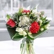 A contemporary vase arrangement of red and white flowers stands in soft daylight, like a cool breath of fresh air drifting in from the Thames at Nine Elms. At the heart of the bouquet, a single coral-red rose opens in full bloom, its velvety petals curling outward with quiet confidence. Beside it, a ruffled crimson carnation adds drama and depth, its layered petals creating a sense of movement and emotion. Around these bold focal blooms, clusters of tiny white daisy-like flowers and neat button chrysanthemums scatter light through the design, their crisp petals echoing the clean brightness of the room. Creamy white lily buds rise above and between the other flowers, still closed, their long tapered forms suggesting future fragrance and unfolding beauty. Broad, glossy green leaves sweep around the base in graceful arcs, almost sculptural, cradling the stems and giving the whole arrangement a polished, architectural silhouette. The flowers sit in a clear glass vase, the stems visible through the water, with reflections of the window washing over the surface. The lighting suggests a bright yet calm London morning, like the gentle glow along the new streets of Nine Elms, and you can almost sense a faint green, slightly sweet scent on the air. This arrangement feels both modern and timeless, suitable for celebrating a special milestone, offering congratulations in a new Battersea Power Station apartment, or simply bringing a little serenity to a kitchen table overlooking the river.